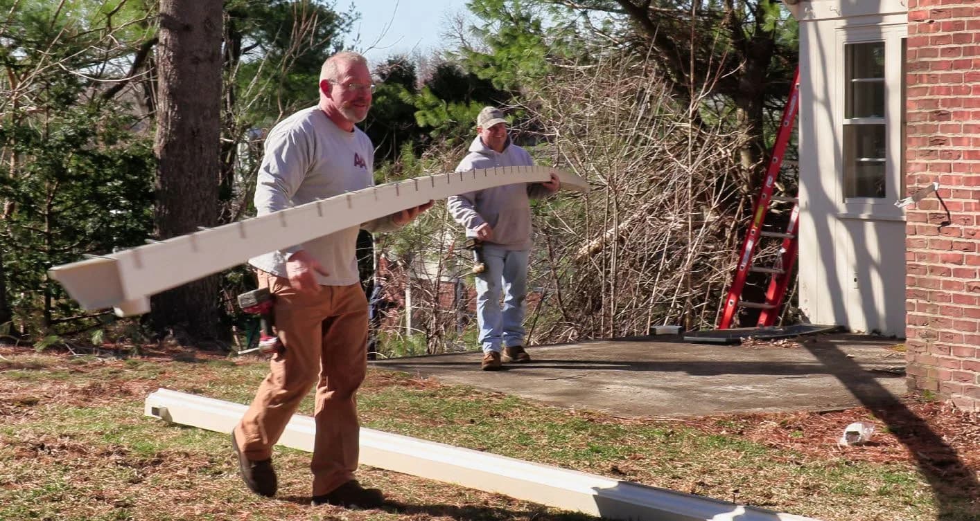 Two workers carrying long gutter section toward brick house
