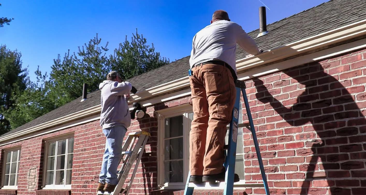 Two A&A workers on ladders installing gutters on brick ranch house